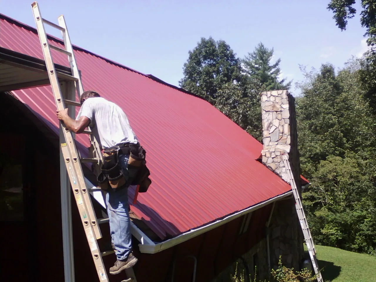 Roofer on a ladder beside a red metal roof and stone chimney in Chestnut Ridge